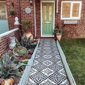 May include: Exterior view of a house with a red brick facade, a green front door, and a pathway adorned with black and white patterned tiles. The pathway is edged with a white picket fence and surrounded by plants, creating a charming entrance.