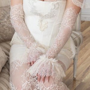 May include: Ivory lace wedding dress with a sweetheart neckline and delicate beaded embellishments. The dress features long, sheer lace sleeves and a matching lace skirt. A sheer lace veil is also visible.