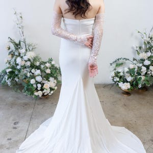 May include: A floor-length white strapless gown featuring a smooth, fitted bodice and a flared skirt with a train. The garment is paired with long, sheer white lace sleeves that extend from the upper arms to the wrists, showcasing intricate floral patterns.