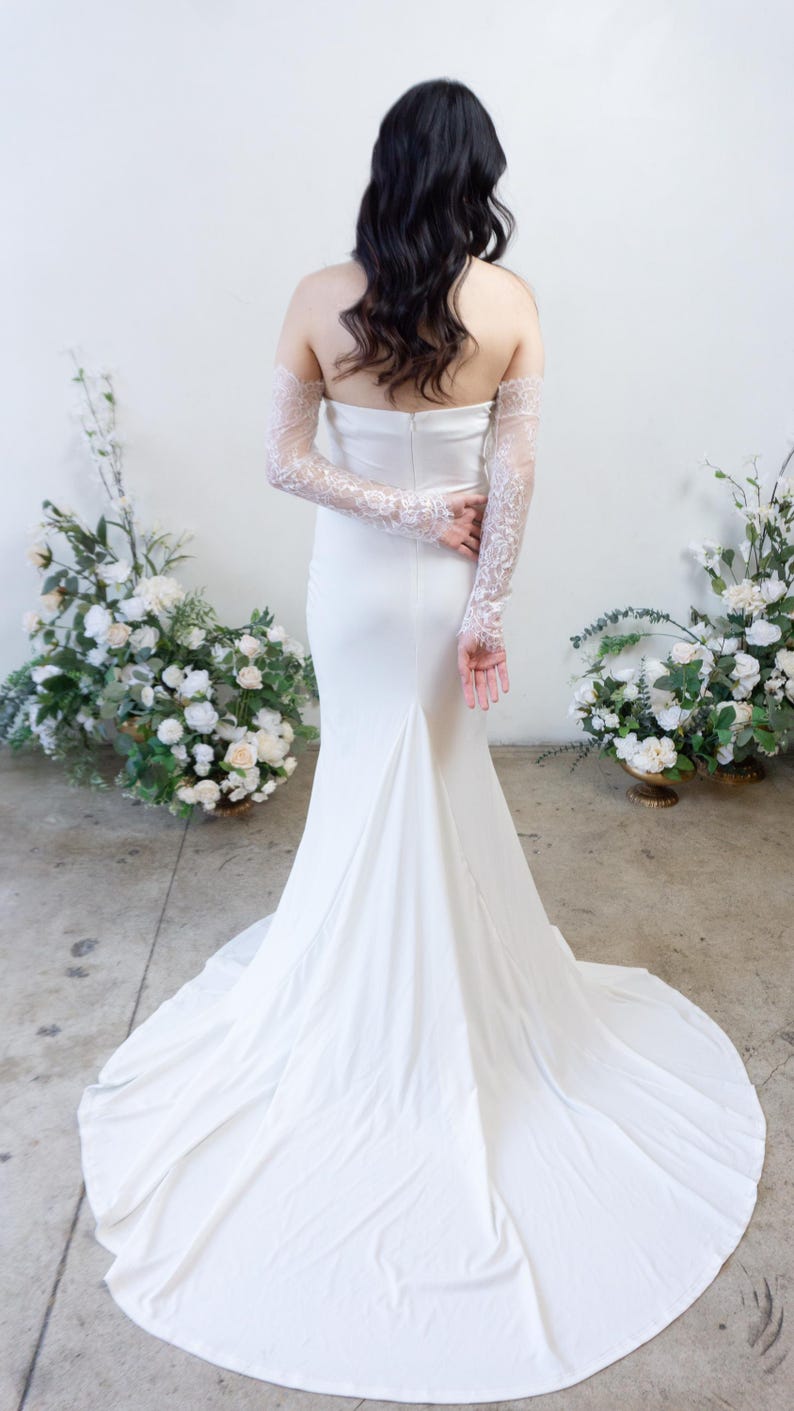 May include: A floor-length white strapless gown featuring a smooth, fitted bodice and a flared skirt with a train. The garment is paired with long, sheer white lace sleeves that extend from the upper arms to the wrists, showcasing intricate floral patterns.