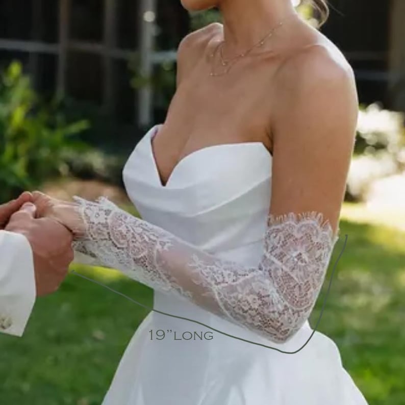 May include: A pair of white lace sleeves featuring a floral pattern and scalloped edges. The sleeves are 19 inches long and are worn with a strapless white gown. The lace fabric extends from the wrist to the upper arm, showcasing intricate detailing.