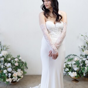 May include: A floor-length white strapless gown featuring a structured corset-style bodice with vertical seams and a fitted silhouette that flares slightly at the hem. The dress is paired with sheer, elbow-length lace sleeves with floral patterns.