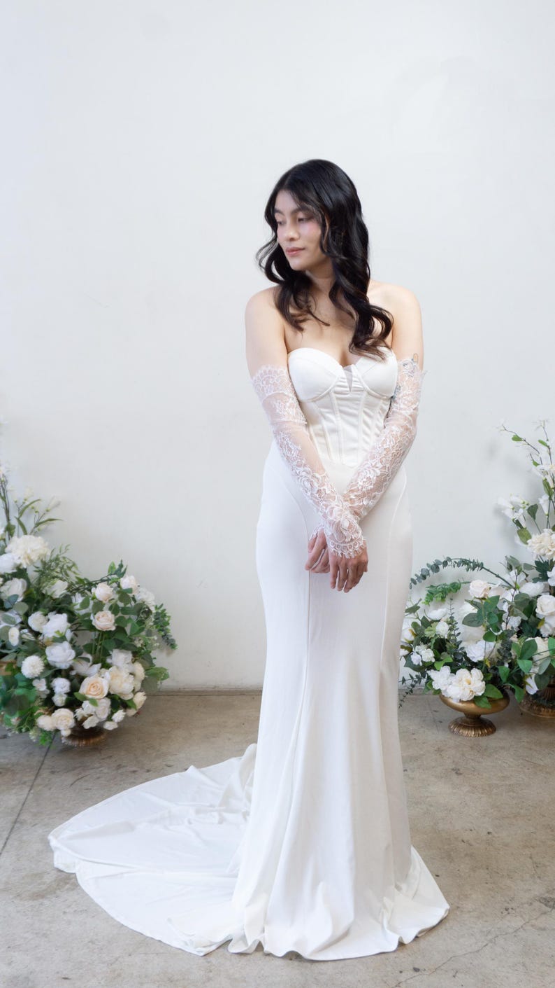 May include: A floor-length white strapless gown featuring a structured corset-style bodice with vertical seams and a fitted silhouette that flares slightly at the hem. The dress is paired with sheer, elbow-length lace sleeves with floral patterns.