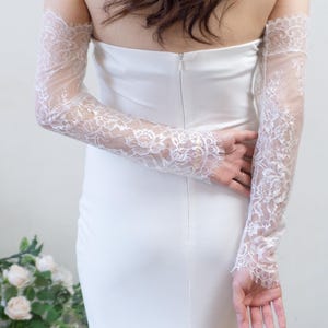 May include: A strapless white floor-length gown featuring a fitted silhouette and a center back zipper, paired with detachable long sleeves made of sheer white floral lace with scalloped edges that extend from the upper arms to the wrists.