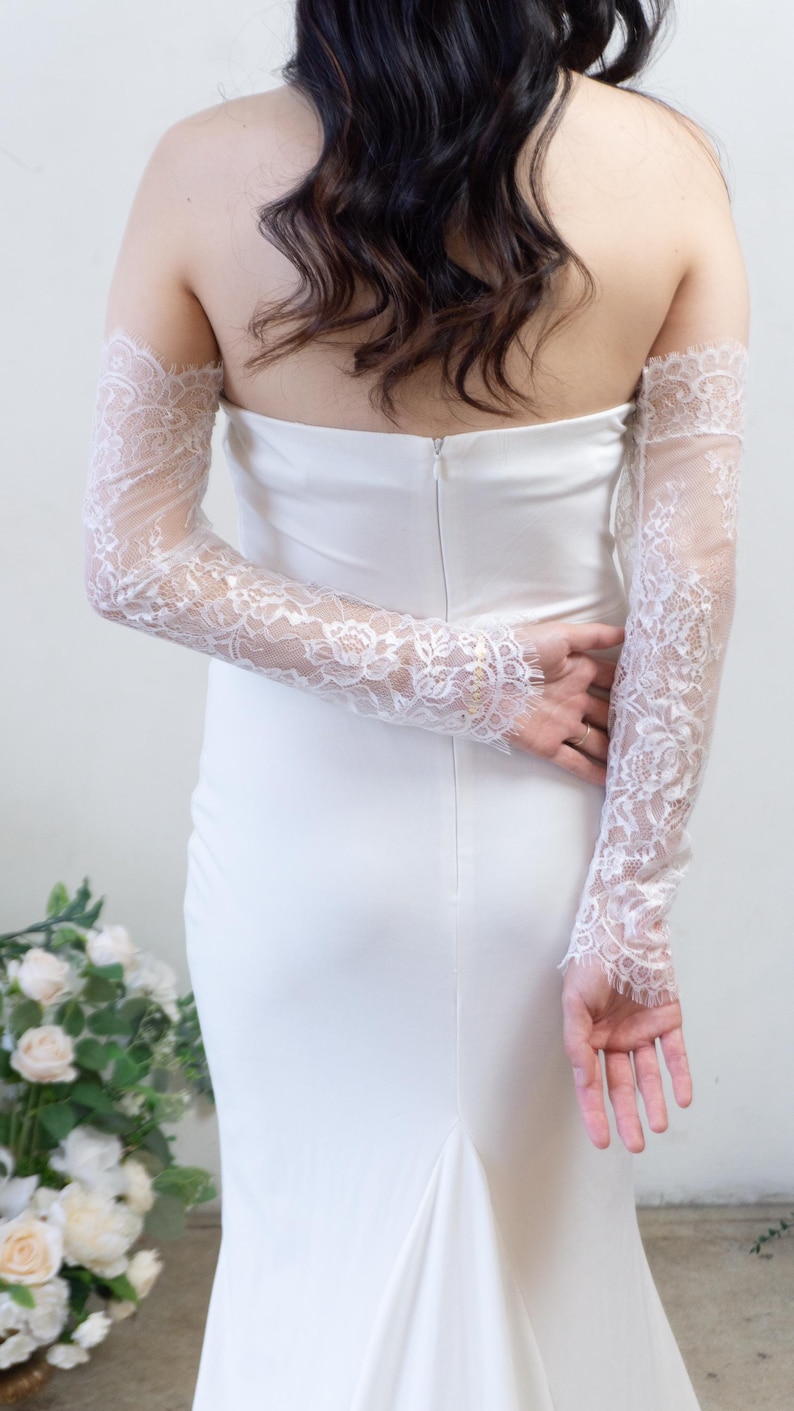 May include: A strapless white floor-length gown featuring a fitted silhouette and a center back zipper, paired with detachable long sleeves made of sheer white floral lace with scalloped edges that extend from the upper arms to the wrists.
