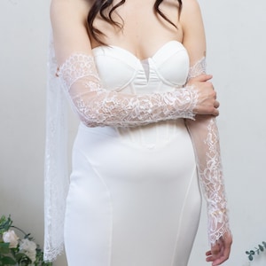 May include: A white strapless corset-style dress featuring a sweetheart neckline and a smooth, fitted bodice. The garment is paired with sheer, long lace sleeves that feature intricate floral patterns and scalloped edges, along with a matching lace scarf.