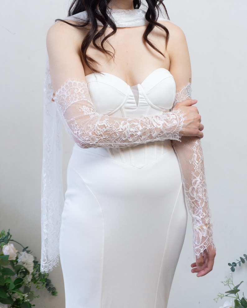 May include: A white strapless corset-style dress featuring a sweetheart neckline and a smooth, fitted bodice. The garment is paired with sheer, long lace sleeves that feature intricate floral patterns and scalloped edges, along with a matching lace scarf.