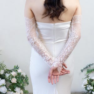 May include: A strapless white gown featuring a fitted bodice with a central back zipper, paired with long, sheer, off-the-shoulder lace sleeves that extend to the wrists and feature intricate floral embroidery and scalloped edges at the cuffs.