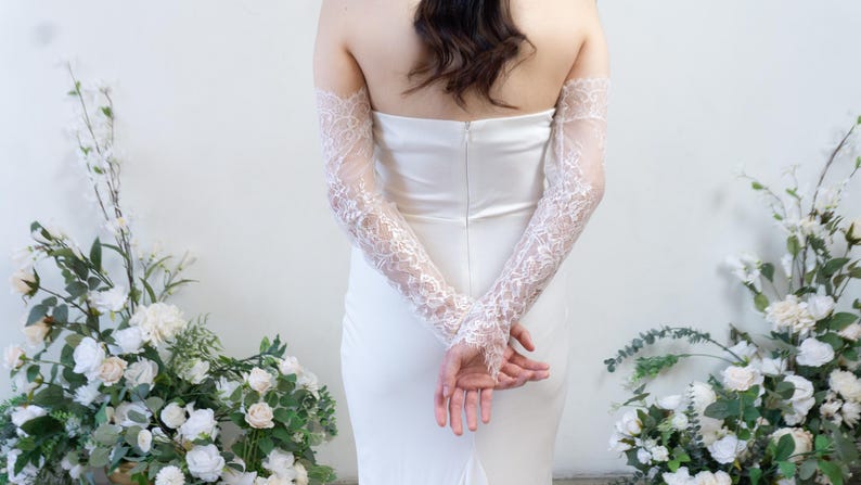 May include: A strapless white gown featuring a fitted bodice with a central back zipper, paired with long, sheer, off-the-shoulder lace sleeves that extend to the wrists and feature intricate floral embroidery and scalloped edges at the cuffs.