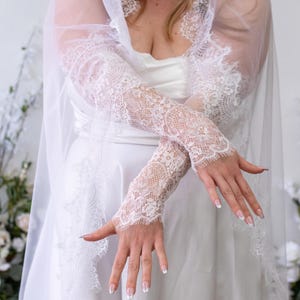 May include: A pair of white lace fingerless gloves featuring intricate floral patterns and scalloped edges, paired with a matching sheer white veil that has a wide lace border. The lace fabric is delicate and semi-sheer, covering the forearms.