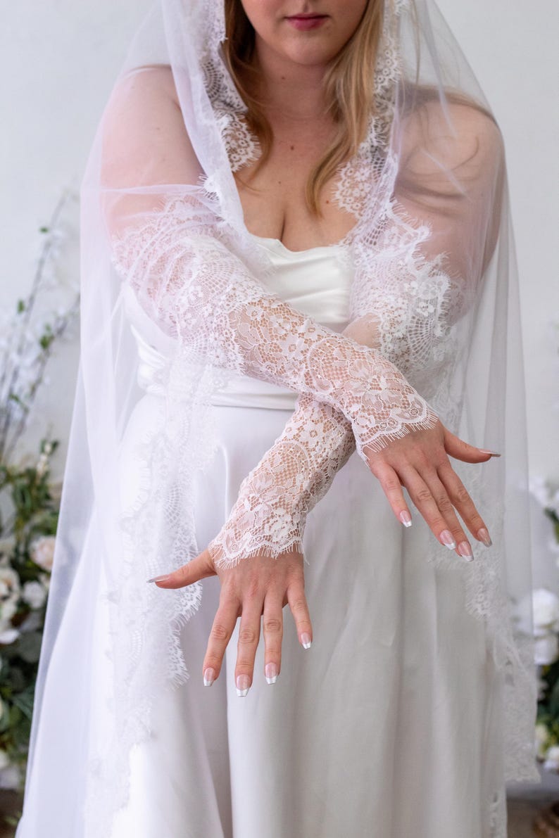 May include: A pair of white lace fingerless gloves featuring intricate floral patterns and scalloped edges, paired with a matching sheer white veil that has a wide lace border. The lace fabric is delicate and semi-sheer, covering the forearms.