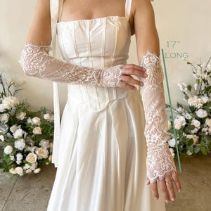 May include: A pair of white lace gloves featuring intricate floral patterns and scalloped edges, measuring 17 inches long. The gloves are worn with a white corset top with ribbon shoulder ties and a matching floor-length skirt.