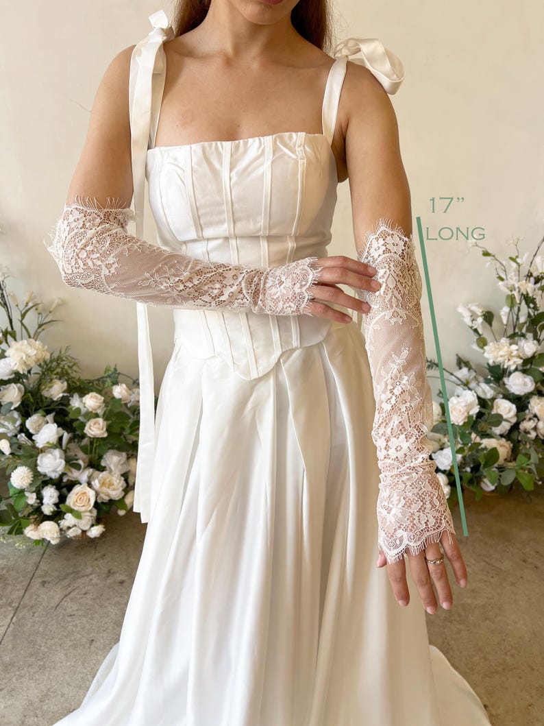 May include: A pair of white lace gloves featuring intricate floral patterns and scalloped edges, measuring 17 inches long. The gloves are worn with a white corset top with ribbon shoulder ties and a matching floor-length skirt.