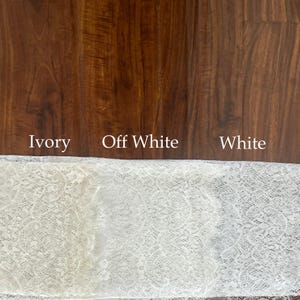 May include: A strip of lace fabric displayed in three distinct color variations labeled Ivory, Off White, and White. The lace features a detailed floral pattern with scalloped edges, showcasing the subtle differences in shade across the fabric.