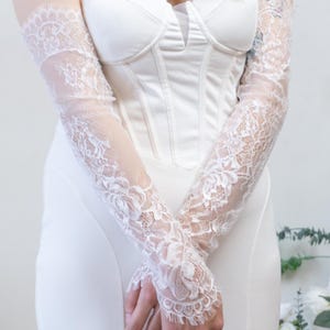 May include: A pair of white lace opera-length gloves featuring a delicate floral pattern and scalloped edges, worn with a strapless white corset-style bodice dress that has a structured sweetheart neckline and a smooth, solid fabric finish.