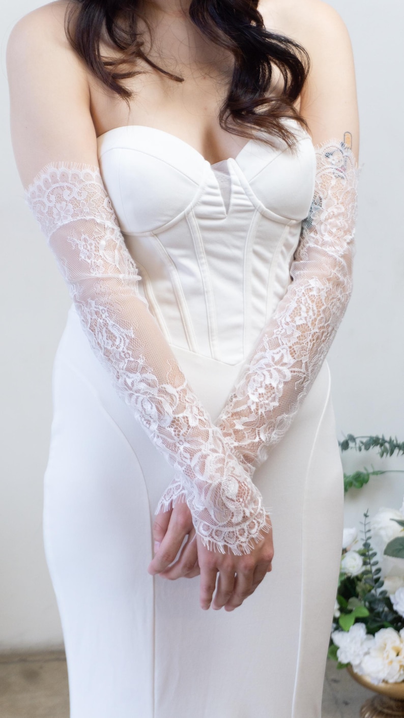May include: A pair of white lace opera-length gloves featuring a delicate floral pattern and scalloped edges, worn with a strapless white corset-style bodice dress that has a structured sweetheart neckline and a smooth, solid fabric finish.