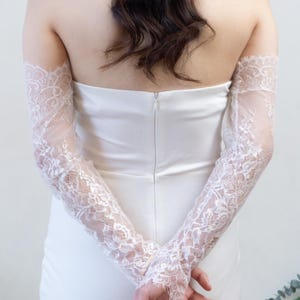 May include: A pair of long, white lace sleeves featuring a delicate floral pattern and scalloped edges. The sleeves are worn over a strapless white gown, extending from the upper arm down to the wrists, showcasing intricate sheer lace detailing.