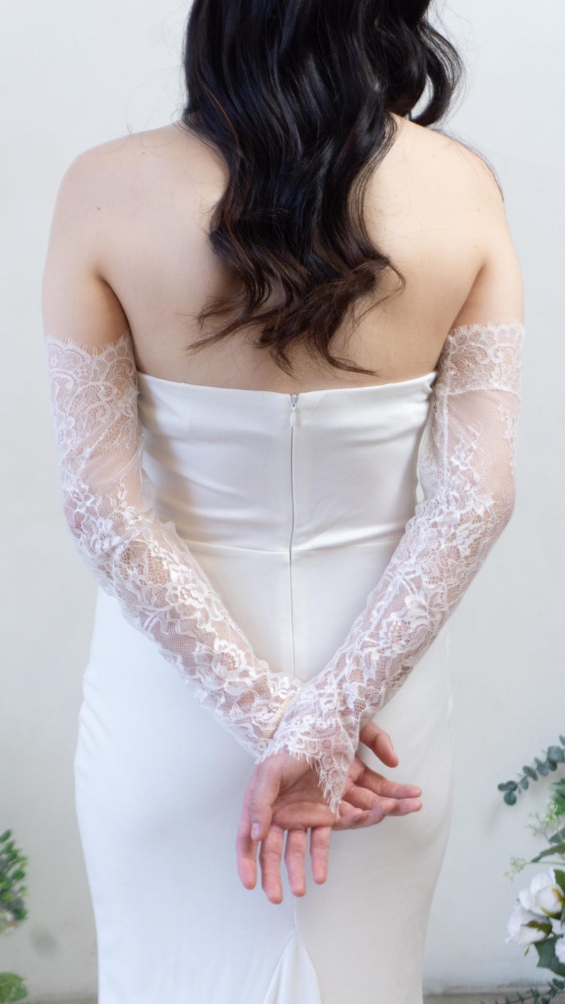 May include: A pair of long, white lace sleeves featuring a delicate floral pattern and scalloped edges. The sleeves are worn over a strapless white gown, extending from the upper arm down to the wrists, showcasing intricate sheer lace detailing.
