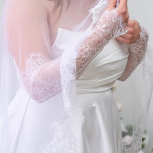 May include: A white strapless dress paired with long, sheer white lace sleeves featuring intricate floral patterns and scalloped edges. A matching white lace-trimmed veil drapes over the shoulders and arms, completing the ensemble.