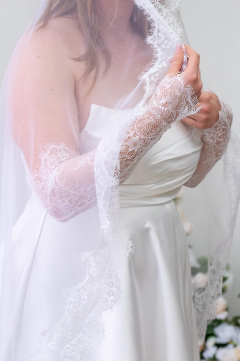 May include: A white strapless dress paired with long, sheer white lace sleeves featuring intricate floral patterns and scalloped edges. A matching white lace-trimmed veil drapes over the shoulders and arms, completing the ensemble.