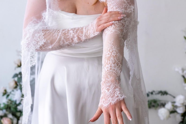 May include: A white satin dress featuring a strapless bodice and long, sheer lace sleeves with intricate floral patterns and scalloped edges. The lace sleeves extend from the shoulders to the wrists, showcasing delicate embroidery and fine mesh detailing.