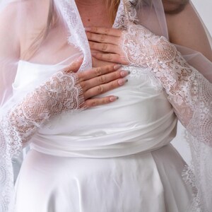 May include: A white strapless dress paired with long, sheer white lace sleeves that extend from the wrists to the upper arms. The lace features a detailed floral pattern with scalloped edges. A matching sheer white veil drapes over the shoulders.