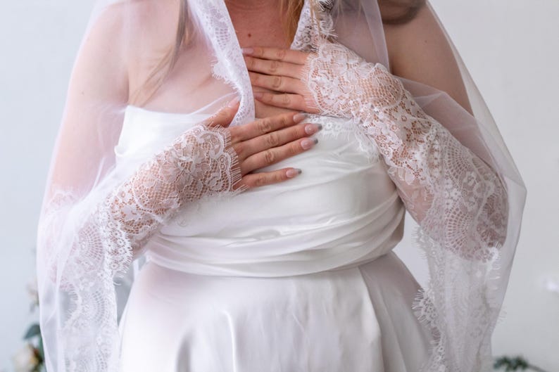May include: A white strapless dress paired with long, sheer white lace sleeves that extend from the wrists to the upper arms. The lace features a detailed floral pattern with scalloped edges. A matching sheer white veil drapes over the shoulders.