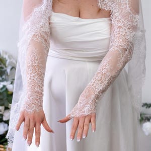 May include: A pair of white lace sleeves featuring intricate floral patterns and scalloped edges, worn with a matching white veil and a strapless white gown. The sleeves extend from the upper arm to the wrist, showcasing delicate lace detailing.