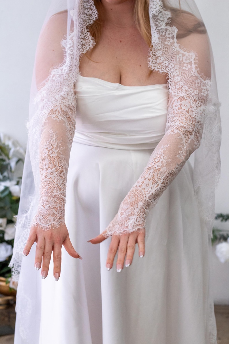 May include: A pair of white lace sleeves featuring intricate floral patterns and scalloped edges, worn with a matching white veil and a strapless white gown. The sleeves extend from the upper arm to the wrist, showcasing delicate lace detailing.