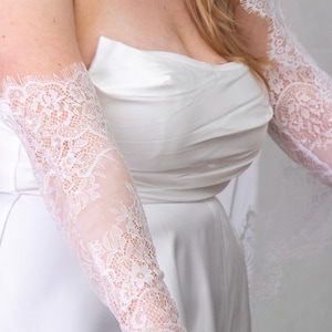 May include: A pair of white lace sleeves featuring a floral pattern with scalloped edges. The sleeves are worn with a strapless white satin dress that has a gathered bodice. The lace extends from the upper arm down to the wrist area.