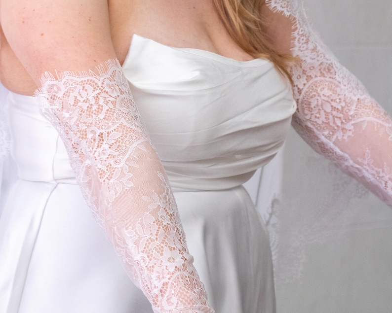 May include: A pair of white lace sleeves featuring a floral pattern with scalloped edges. The sleeves are worn with a strapless white satin dress that has a gathered bodice. The lace extends from the upper arm down to the wrist area.