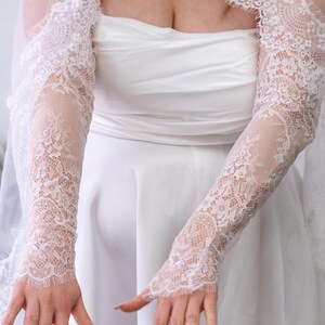 May include: A pair of white lace sleeves featuring a detailed floral pattern and scalloped edges. The sleeves extend from the upper arm to the wrist, covering the arms with sheer, intricate lace fabric that complements the white strapless dress.