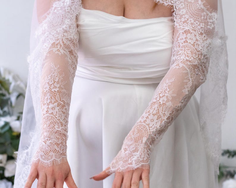 May include: A pair of white lace sleeves featuring a detailed floral pattern and scalloped edges. The sleeves extend from the upper arm to the wrist, covering the arms with sheer, intricate lace fabric that complements the white strapless dress.