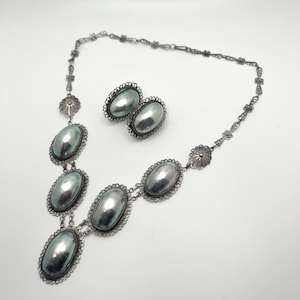 May include: A jewelry set consisting of a necklace and a pair of matching earrings. Each piece features oval, metallic gray cabochons set in intricate, silver-toned filigree metal frames. The necklace chain is composed of delicate, repeating floral links.