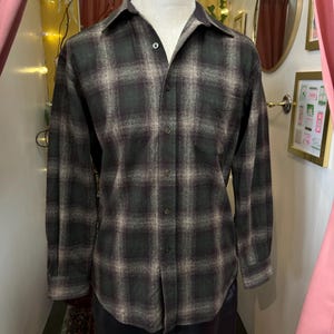 May include: A long-sleeve button-down shirt featuring a dark green and charcoal gray plaid pattern. The shirt has a standard collar, a single chest pocket on the left side, and a full-length front button closure with matching dark buttons.