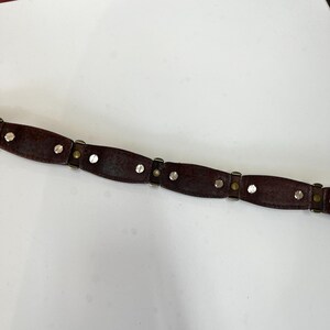 May include: A dark brown leather belt with silver-colored rivets and metal accents. The belt is segmented into oval-shaped sections, connected by smaller metal pieces. The rivets are evenly spaced along each section, creating a decorative pattern.