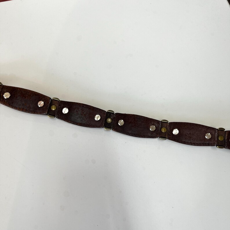 May include: A dark brown leather belt with silver-colored rivets and metal accents. The belt is segmented into oval-shaped sections, connected by smaller metal pieces. The rivets are evenly spaced along each section, creating a decorative pattern.