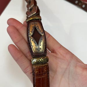 May include: Brown leather belt with ornate metal accents. The belt features a braided design at the top, a decorative center piece with a diamond-shaped cutout, and a textured, crocodile-like pattern on the lower portion.