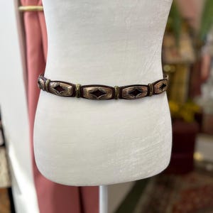 May include: A brown belt with rectangular metal accents and a diamond-shaped design. The belt is displayed on a white mannequin. The metal accents have a silver and gold finish. The belt is a fashion accessory.