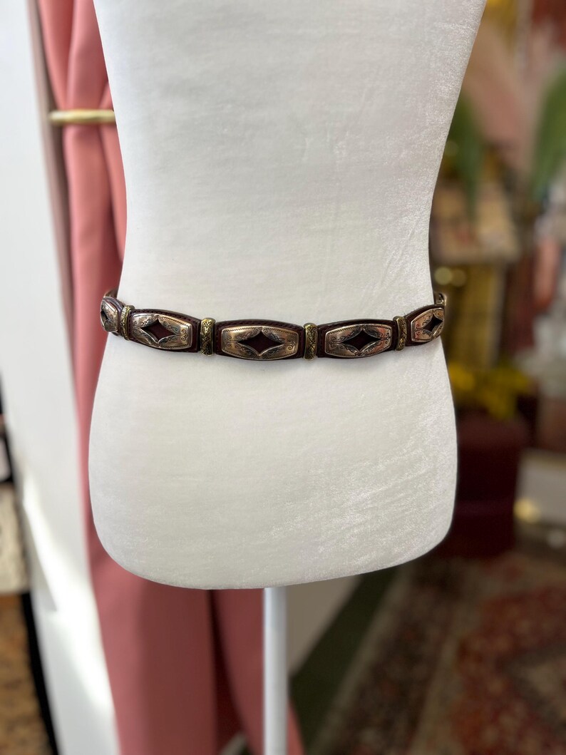 May include: A brown belt with rectangular metal accents and a diamond-shaped design. The belt is displayed on a white mannequin. The metal accents have a silver and gold finish. The belt is a fashion accessory.
