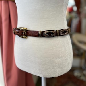 May include: A brown leather belt with a gold-colored buckle and decorative metal accents. The belt features a textured, crocodile-like pattern and rectangular metal embellishments with a copper-colored center.