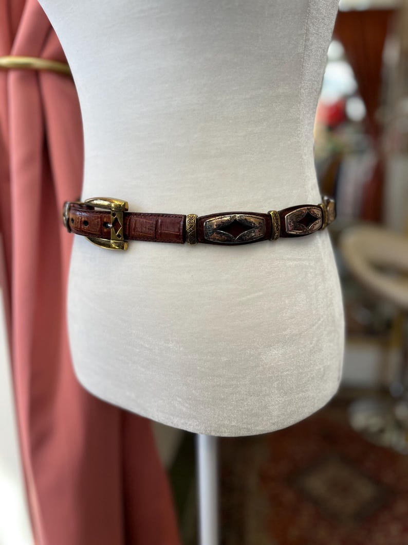 May include: A brown leather belt with a gold-colored buckle and decorative metal accents. The belt features a textured, crocodile-like pattern and rectangular metal embellishments with a copper-colored center.