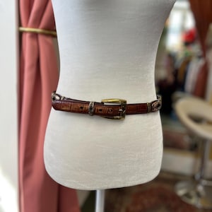 May include: A brown leather belt with a crocodile pattern is displayed on a mannequin. The belt has a gold-colored buckle and decorative metal accents. The belt is approximately 1.5 inches wide.