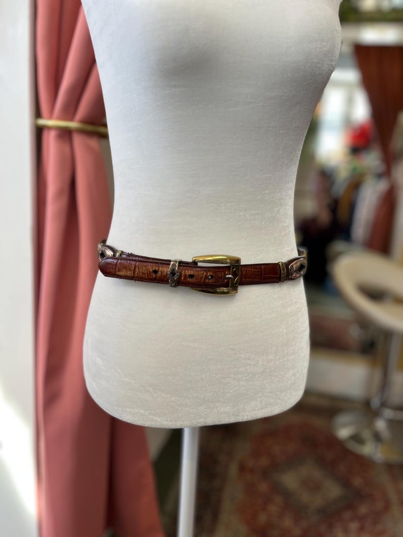 May include: A brown leather belt with a crocodile pattern is displayed on a mannequin. The belt has a gold-colored buckle and decorative metal accents. The belt is approximately 1.5 inches wide.
