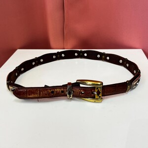 May include: A brown leather belt with a gold-colored buckle and silver-colored accents. The belt has a textured surface and is adorned with small, silver-colored studs. The buckle is rectangular with a decorative design.
