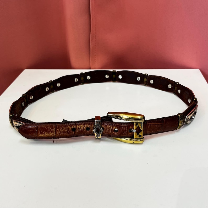 May include: A brown leather belt with a gold-colored buckle and silver-colored accents. The belt has a textured surface and is adorned with small, silver-colored studs. The buckle is rectangular with a decorative design.