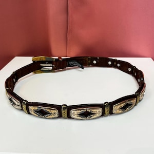 May include: A brown leather belt with decorative metal accents. The belt features rectangular panels with a diamond-shaped design and ornate detailing. The buckle is gold-toned, and the belt has a classic, vintage style.