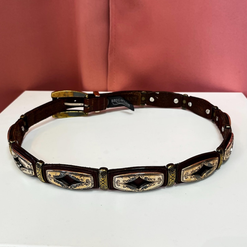 May include: A brown leather belt with decorative metal accents. The belt features rectangular panels with a diamond-shaped design and ornate detailing. The buckle is gold-toned, and the belt has a classic, vintage style.