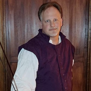 May include: A man wearing a purple vest and white shirt holds a sword in his right hand. The sword has a black leather grip and a silver blade.