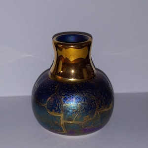 May include: A small, round vase with a dark blue iridescent finish and gold accents. The vase has a gold rim and a pattern of gold lines across the surface. The vase is approximately 4 inches tall.